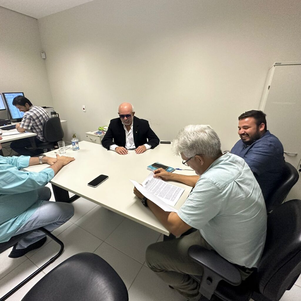 Quatro homens sentados em volta de uma mesa, com papíes para serem assinados, com uma quinta pessoa usando um computador em outra mesinha.