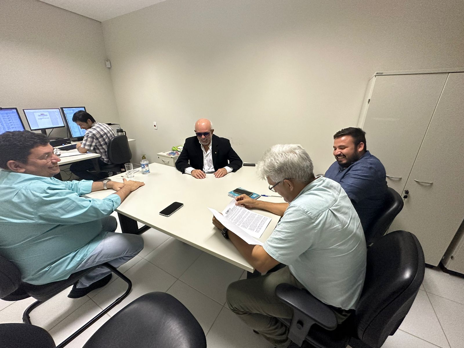 Quatro homens sentados em volta de uma mesa, com papíes para serem assinados, com uma quinta pessoa usando um computador em outra mesinha.