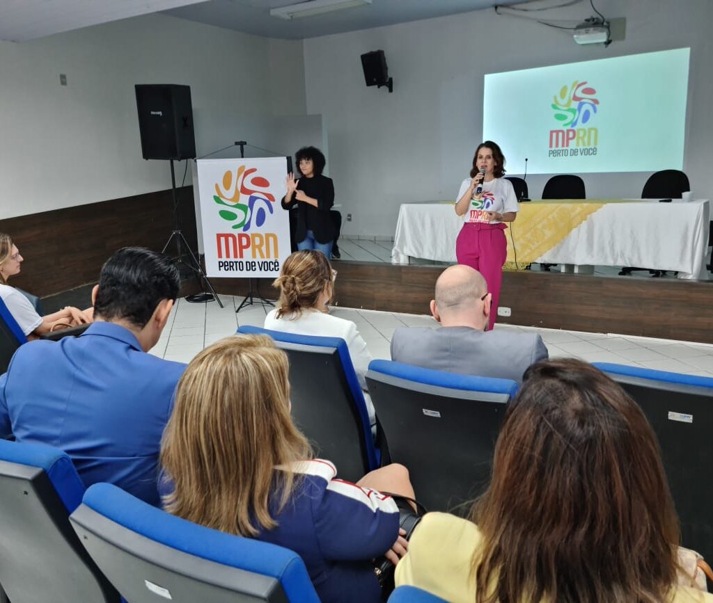 A PGJ, usando calça rosa e camiseta do projeto MPRN perto de você está falando com o microfone na mão para uma plateia sentada nas cadeiras de um auditório.