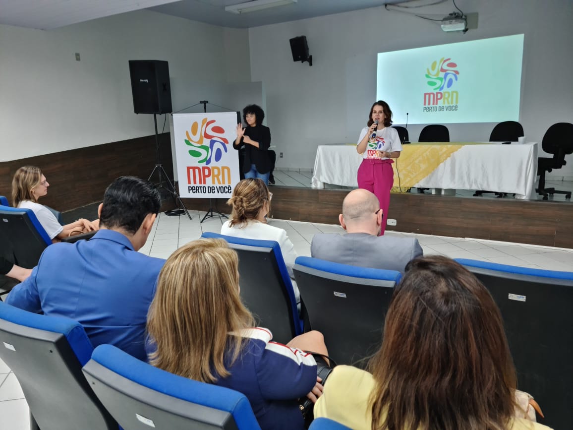 A PGJ, usando calça rosa e camiseta do projeto MPRN perto de você está falando com o microfone na mão para uma plateia sentada nas cadeiras de um auditório.