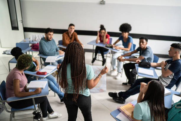 Imagem com várias pessoas sentadas em carteiras escolares, em um círculo, e uma pessoa em pé, segurando um cartaz e falando com elas.