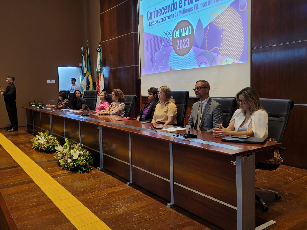 Imagem com várias pessoas sentadas à mesa, em um auditório