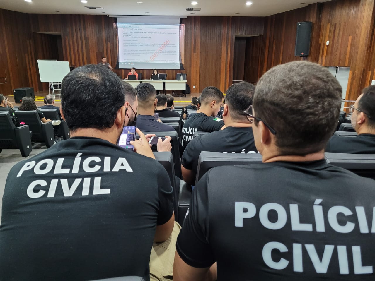 Imagem mostras homensa com camisetas pretas com o nome "Polícia Civil" em branco. Estão sentado em cadeiras de um auditório.