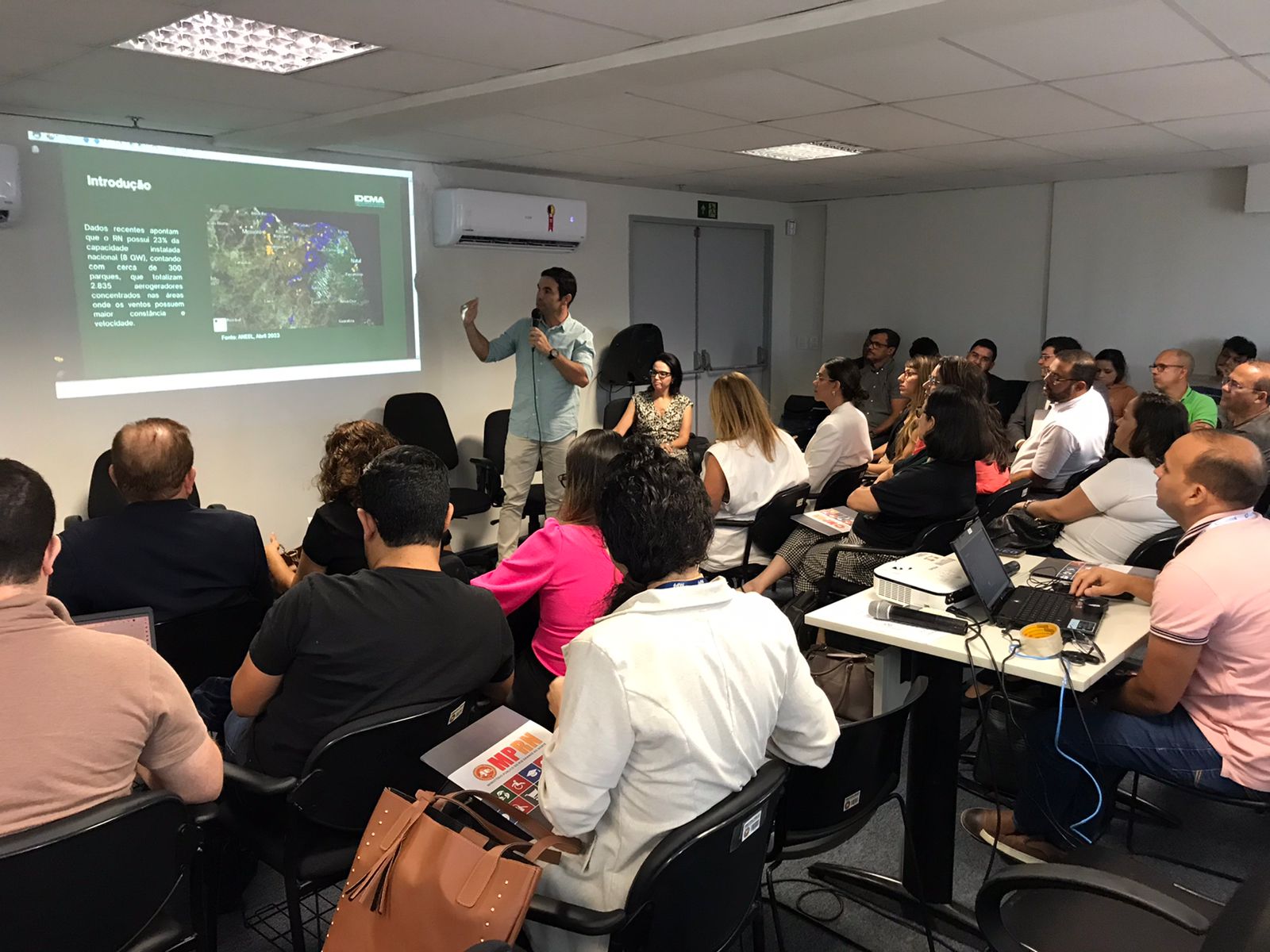 Imagem de um mini-auditório com várias pessoas sentadas, assistindo a uma palestra.