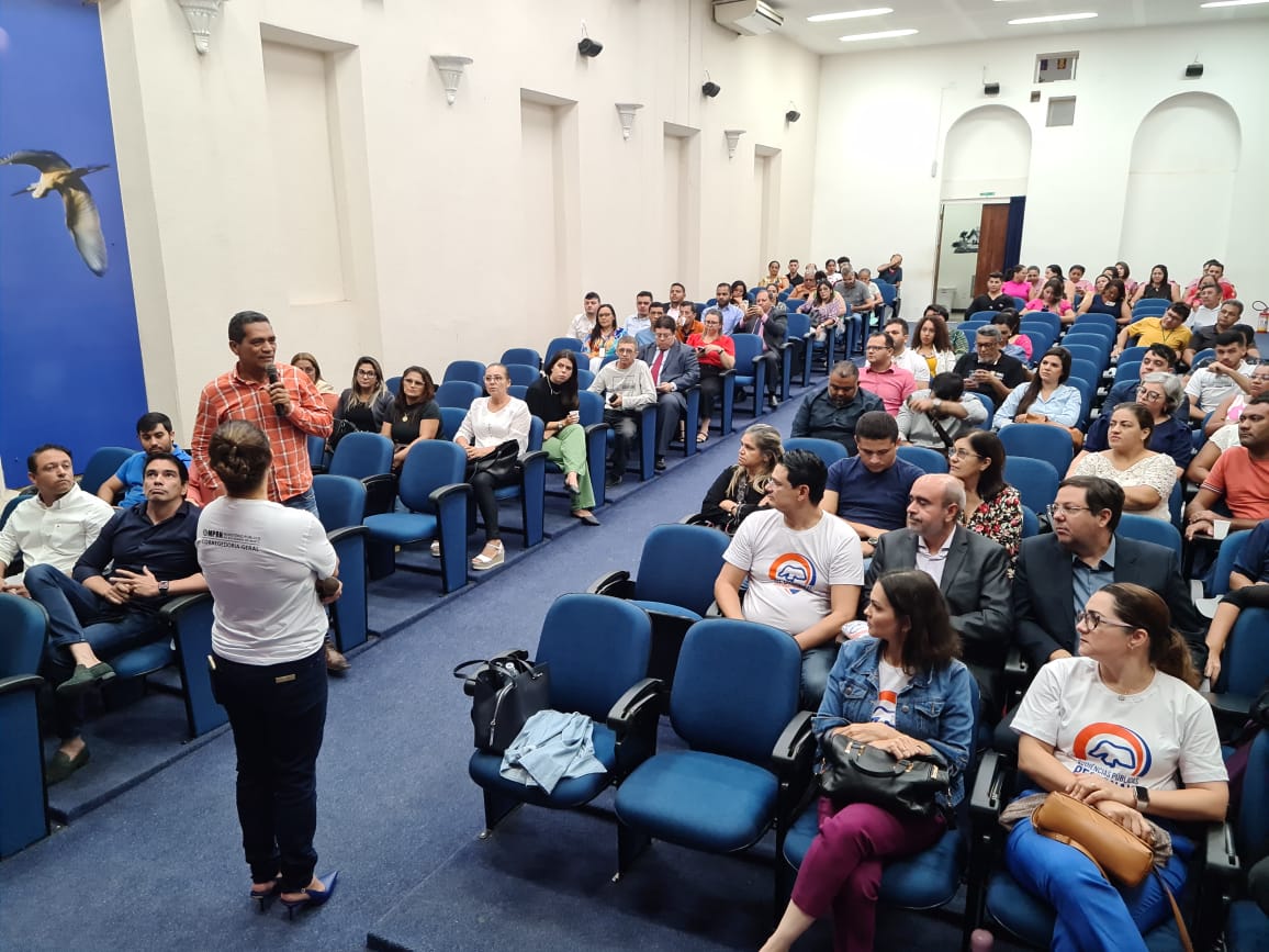 Imagem de um auditório com várias pessoas sentadas e uma mulher ao centro, de frente para a plateia.