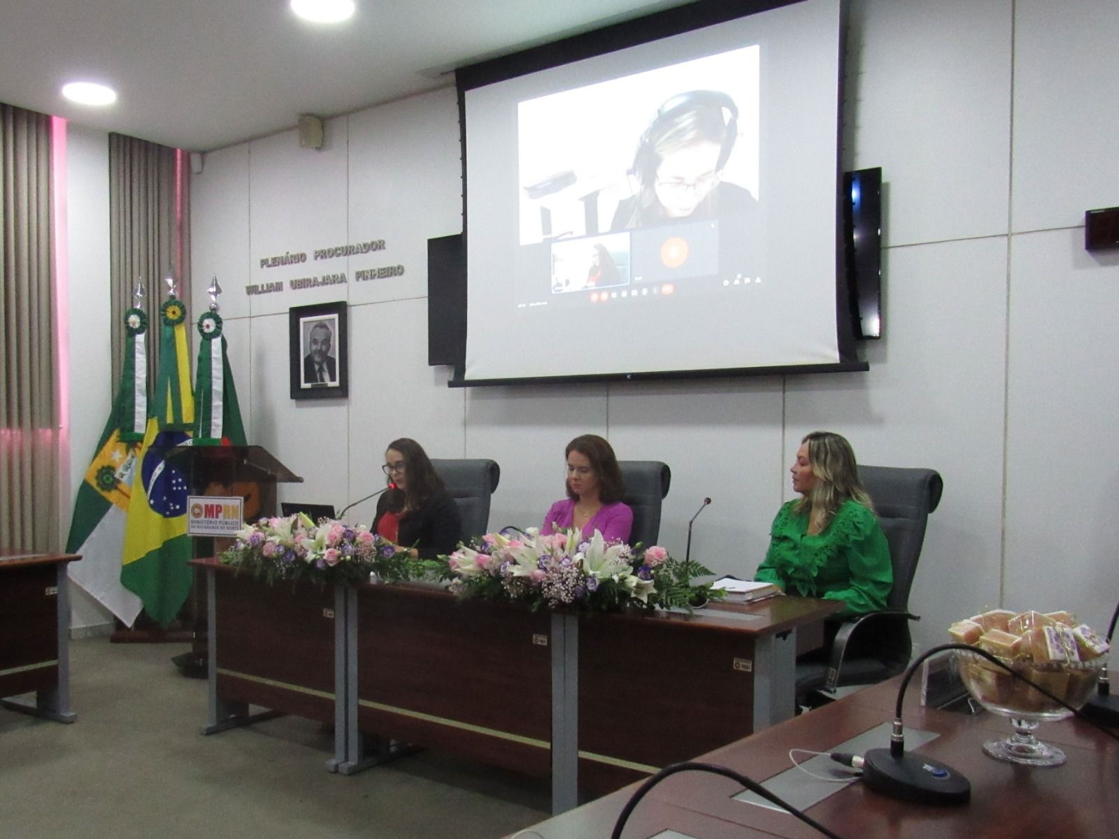 Imagem de três mulheres compondo uma mesa em um plenário, com um telão atrás delas