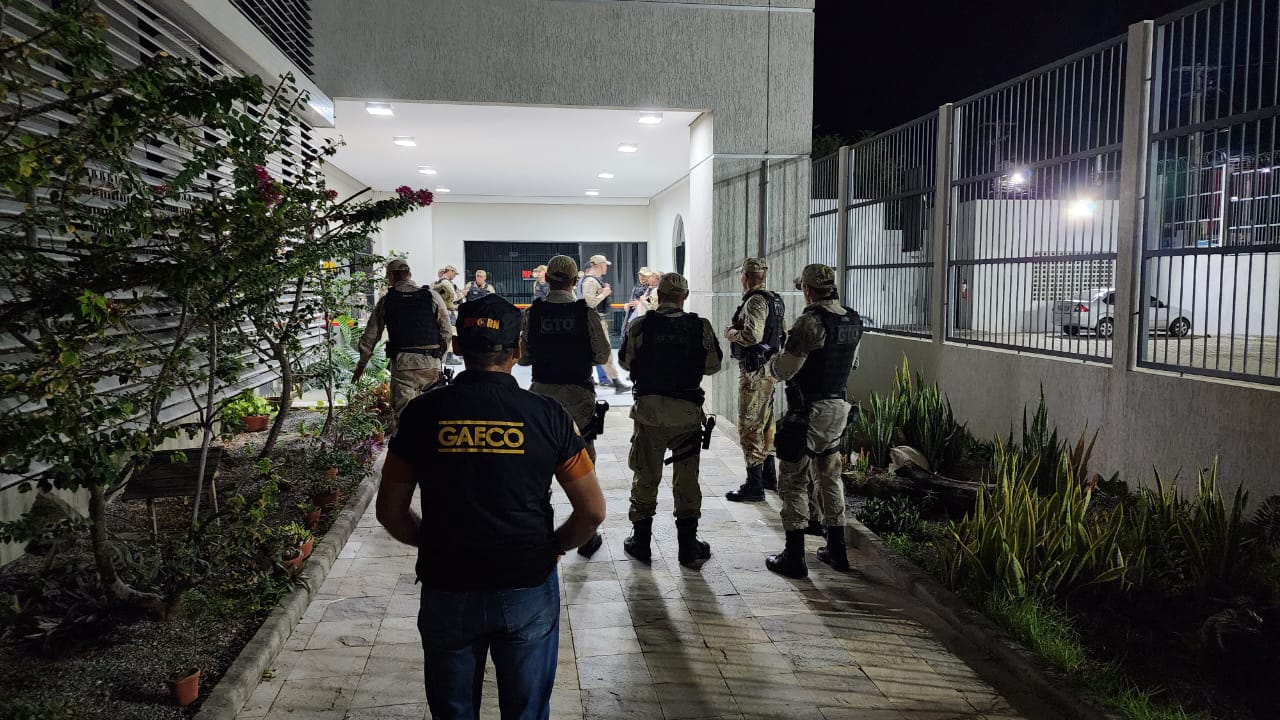 Imagem com um agente do Gaeco e policiais de costas, em frente a um prédio.