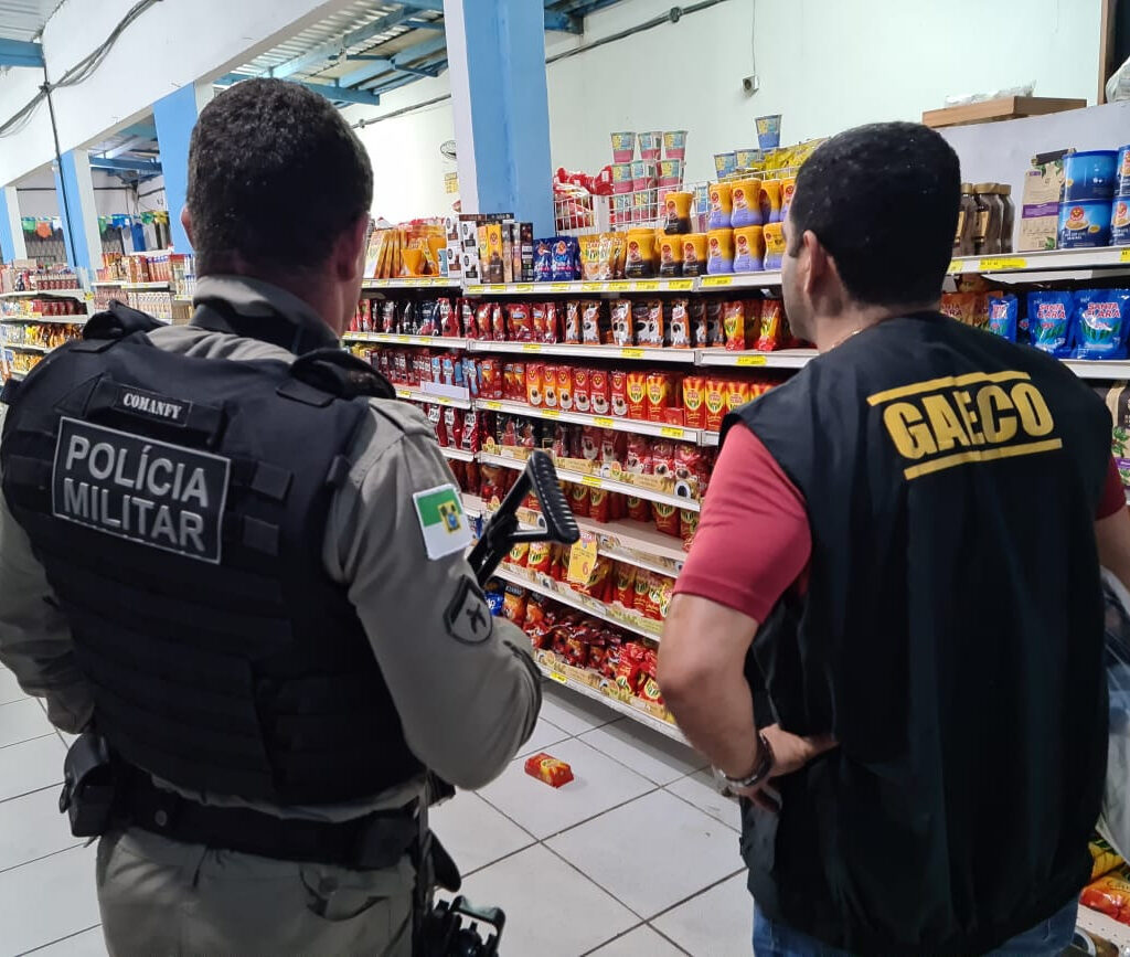 Imagem de um policial militar e uma agente do Gaeco, ambos de costas, em um supermercado.