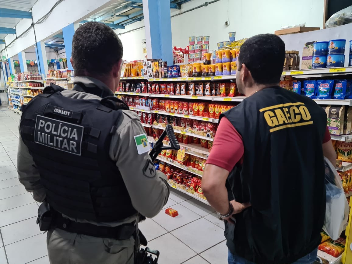 Imagem de um policial militar e uma agente do Gaeco, ambos de costas, em um supermercado.