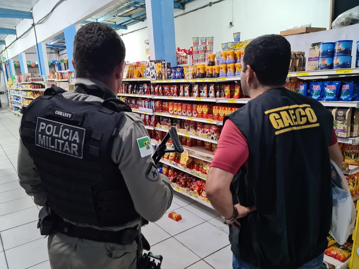 Imagem de um policial militar e uma agente do Gaeco, ambos de costas, em um supermercado.