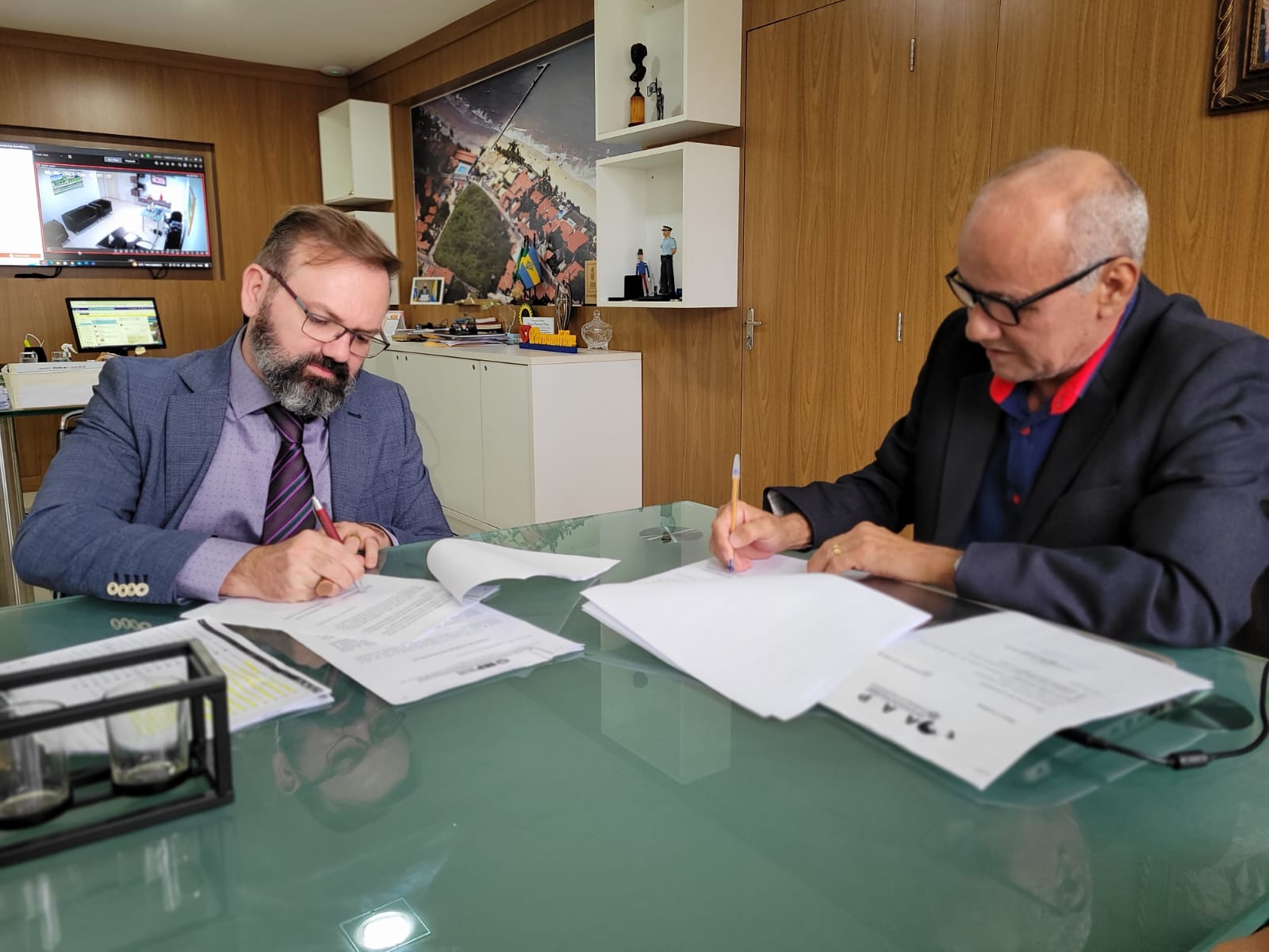 Imagem de dois homens sentados à mesa, cada um assinando um documento.