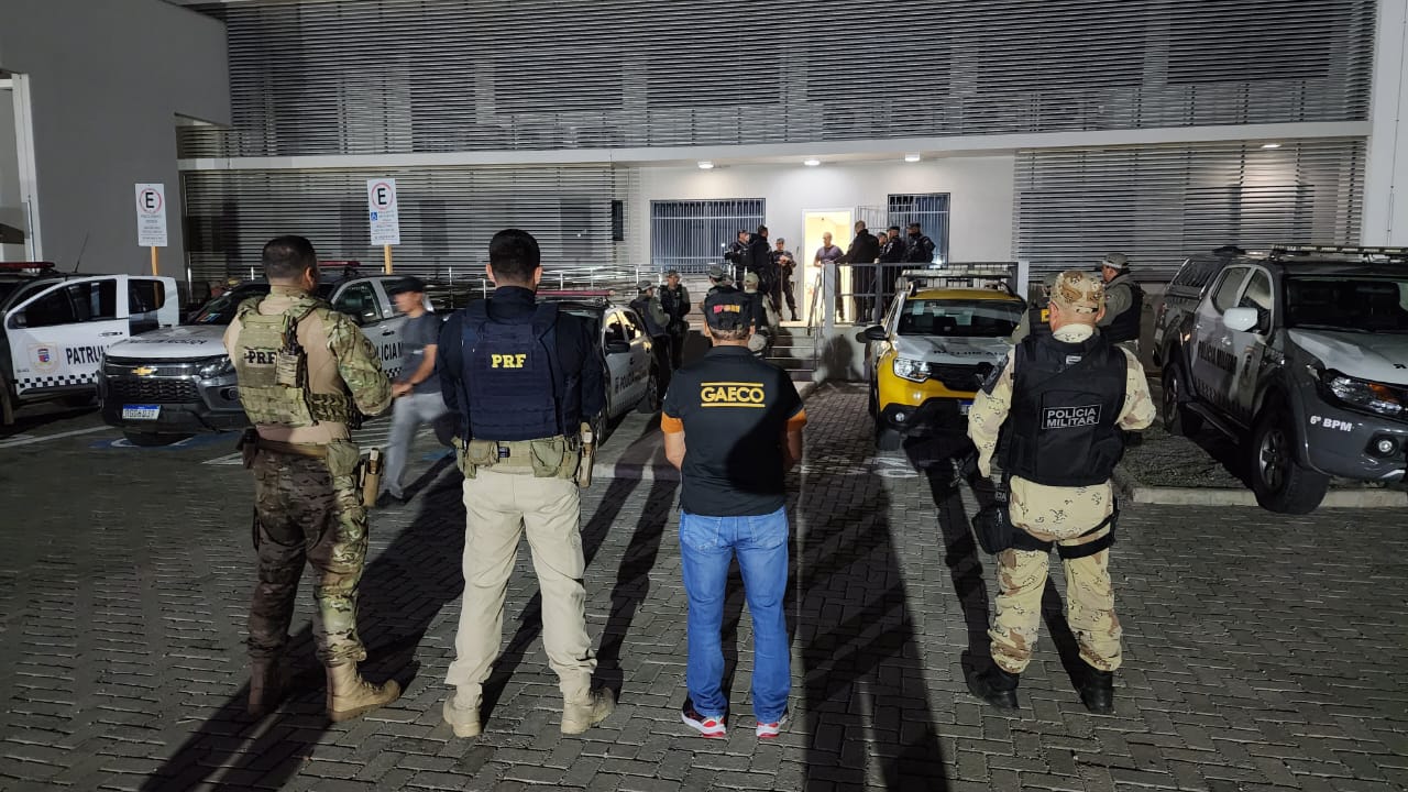 Imagem de agentes do Gaeco e policiais rodoviários federais e militares, de costas, em primeiro plano. Ao fundo, viaturas e mais policiais em frente a um prédio.