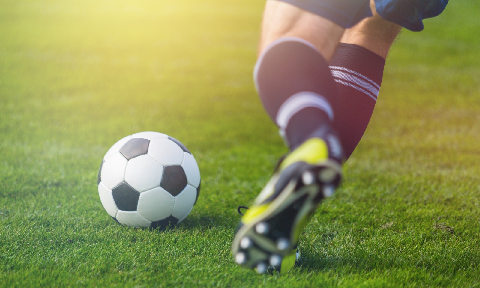 Imagem de um jogador, aparecendo apenas as pernas, correndo atrás de uma bola de futebol.