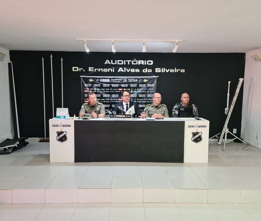 Imagem de um promotor de justiça e três policiais militares sentados à mesa no auditório do estádio Frasqueirão.