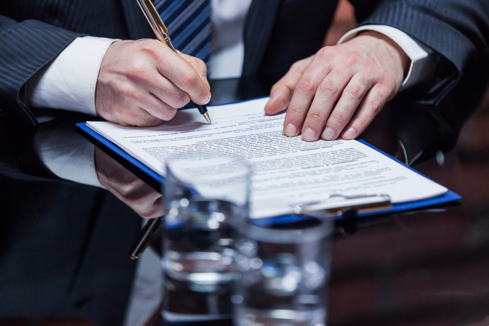 Imagem de um homem, vestido com terno e gravata e sem aparecer o rosto, sentado, assinando um documento que está sobre a mesa.