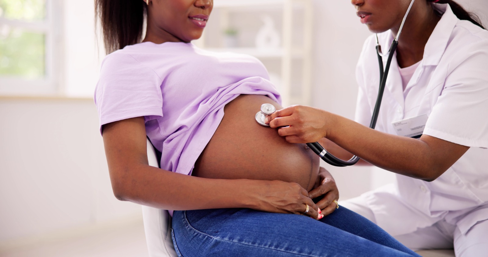 Imagem de uma mulher negra, grávida, sentada em uma cadeira e uma profissional de saúde fazendo a ausculta do bebê.