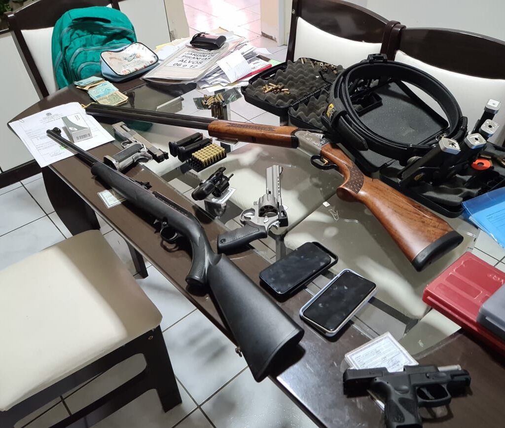 Imagem de várias armas, munições, documentos, aparelhos celulares e dinheiro sobre uma mesa de vidro.
