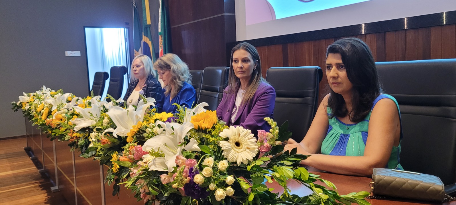 Imagem de quatro mulheres sentadas à mesa, em um auditório.