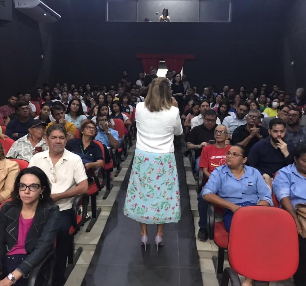 Imagem de uma mulher de costas, falando para uma plateia, em um auditório.