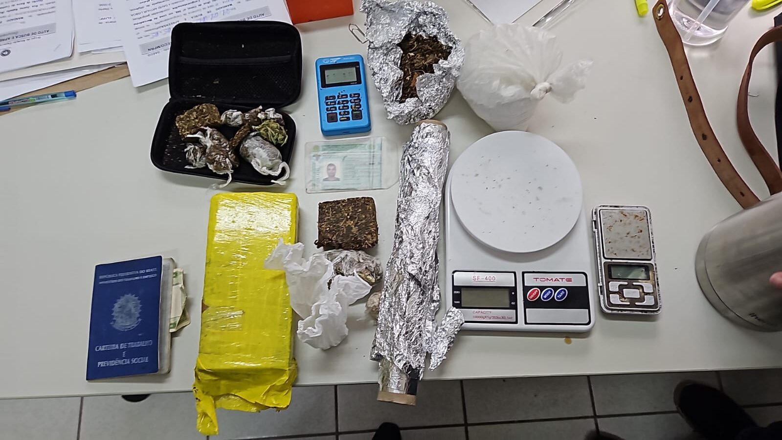 Imagem de uma mesa com pacotes de drogas, balança de precisão, maquinetas de cartão, aparelho celular e documentos.