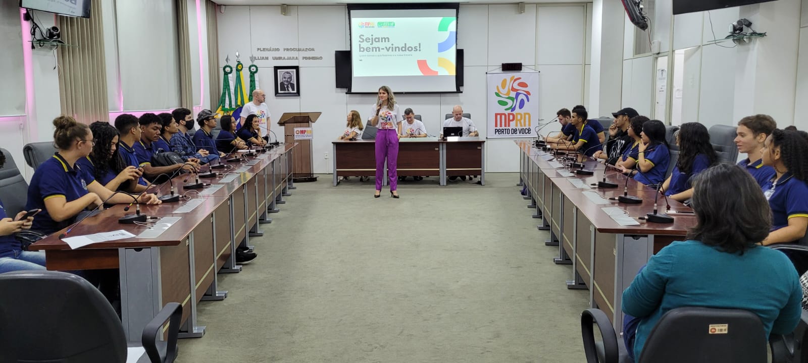 Imagem de um plenário com uma mulher falando ao microfone para estudantes sentados em bancadas.