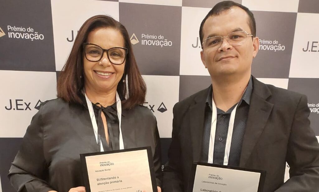 Imagem de uma mulher e um homem, ambos em pé, segurando placas de premiação.