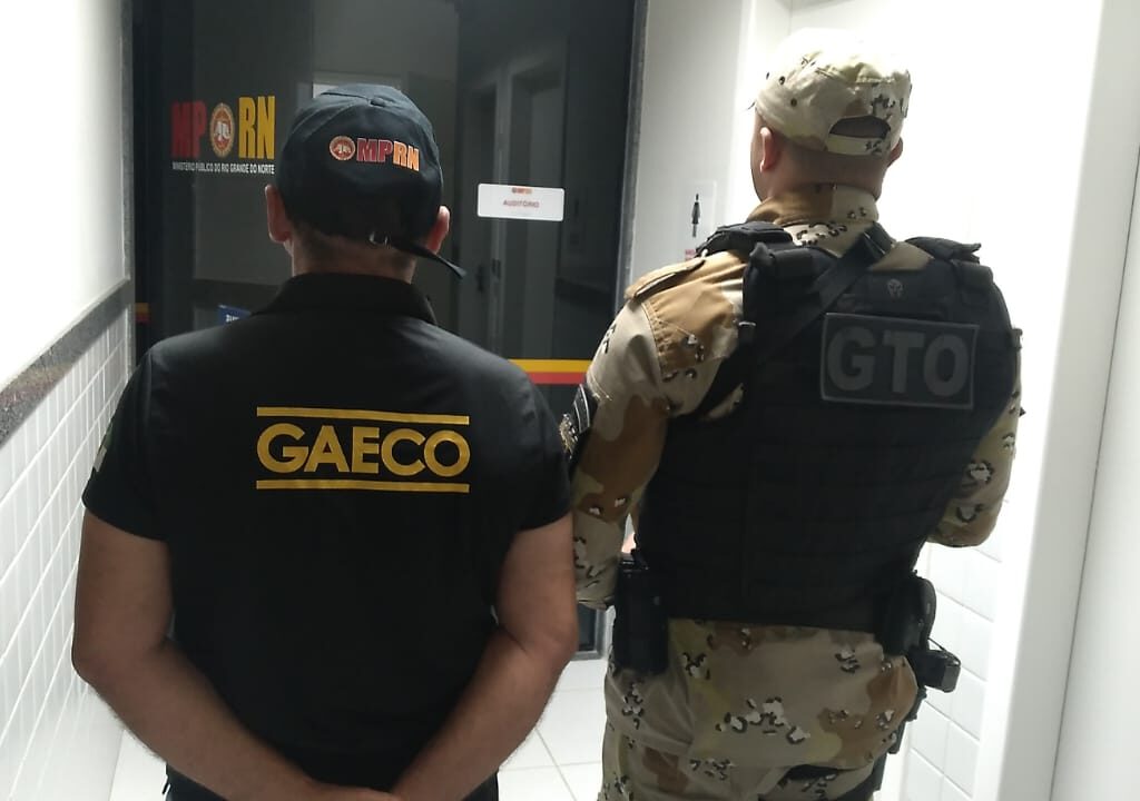 Imagem de um agente do Gaeco e de um policial do GTO, ambos de costas, em pé, em frente a uma porta de vidro.