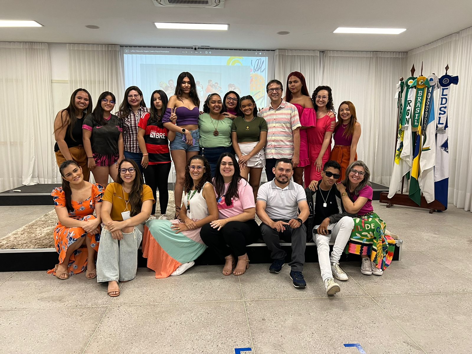 Foto mostra adolescentes e padinhos e madrinhas dispostos em duas fileiras (a da frente, estão sentados no tabalhado do palco e a de trás, estão em pé no tablado).
