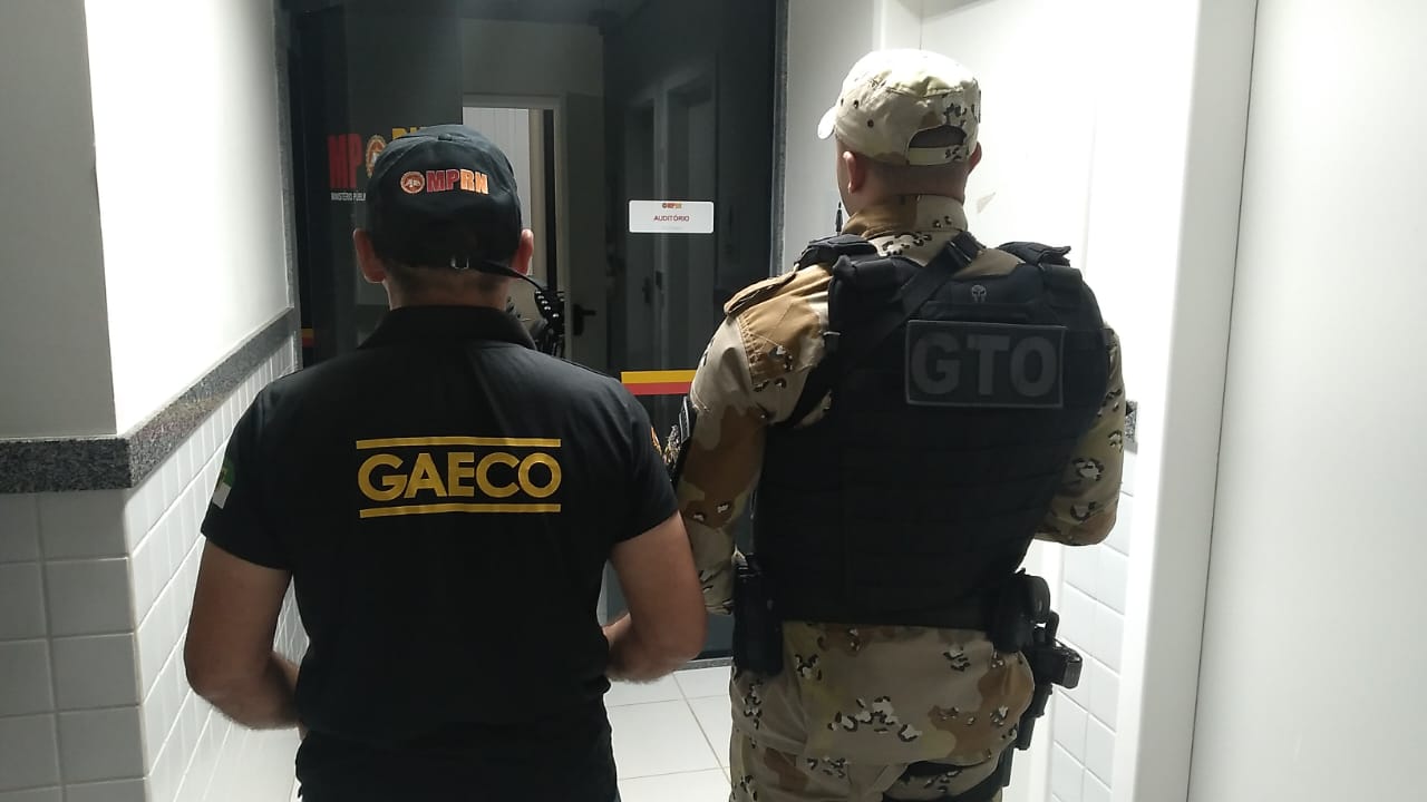 Imagem de um agente do Gaeco e de um policial militar, ambos de costas e em pé, em frente a uma porta de vidro.