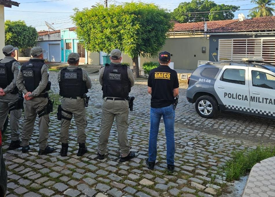 Imagem de quatro policiais militares e um integrante do Gaeco, todos de costas, em pé, em frente a uma rua.