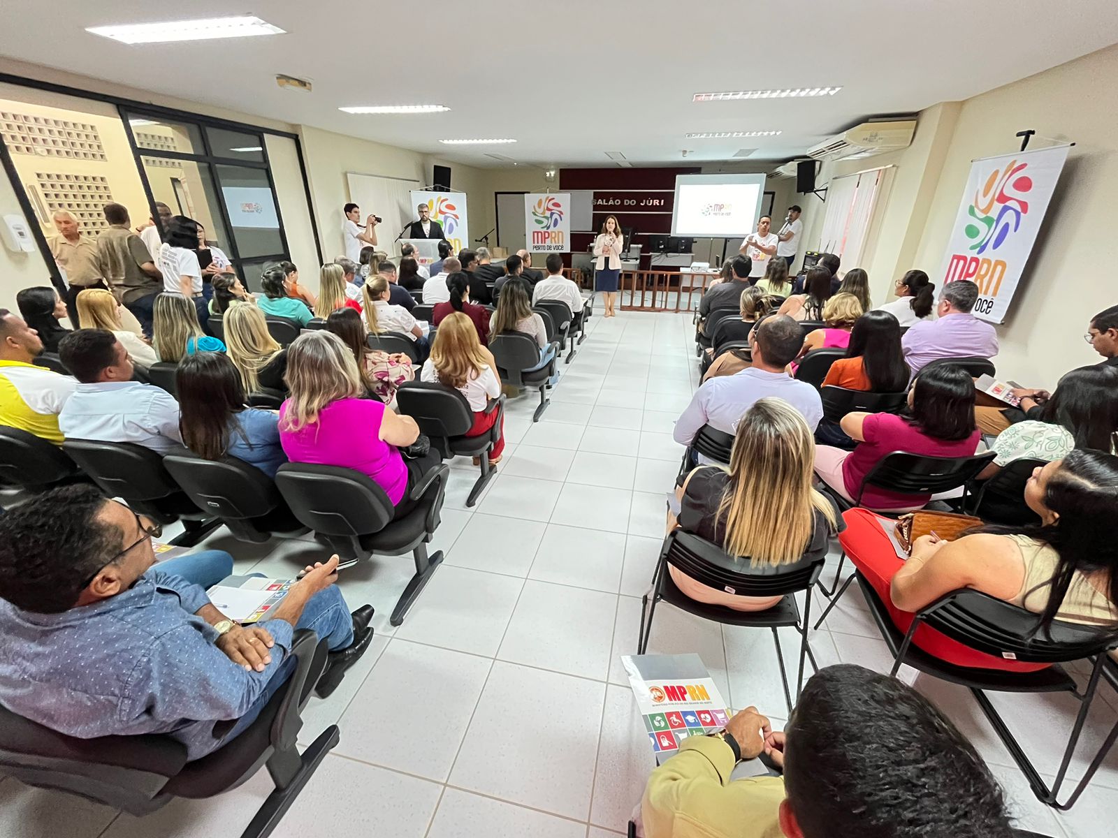 Imagem de várias pessoas sentadas, em um auditório, na abertura do MPRN Perto de Você, na cidade de Santo Antônio, ouvindo a procuradora-geral de Justiça Elaine Cardoso falar ao microfone.
