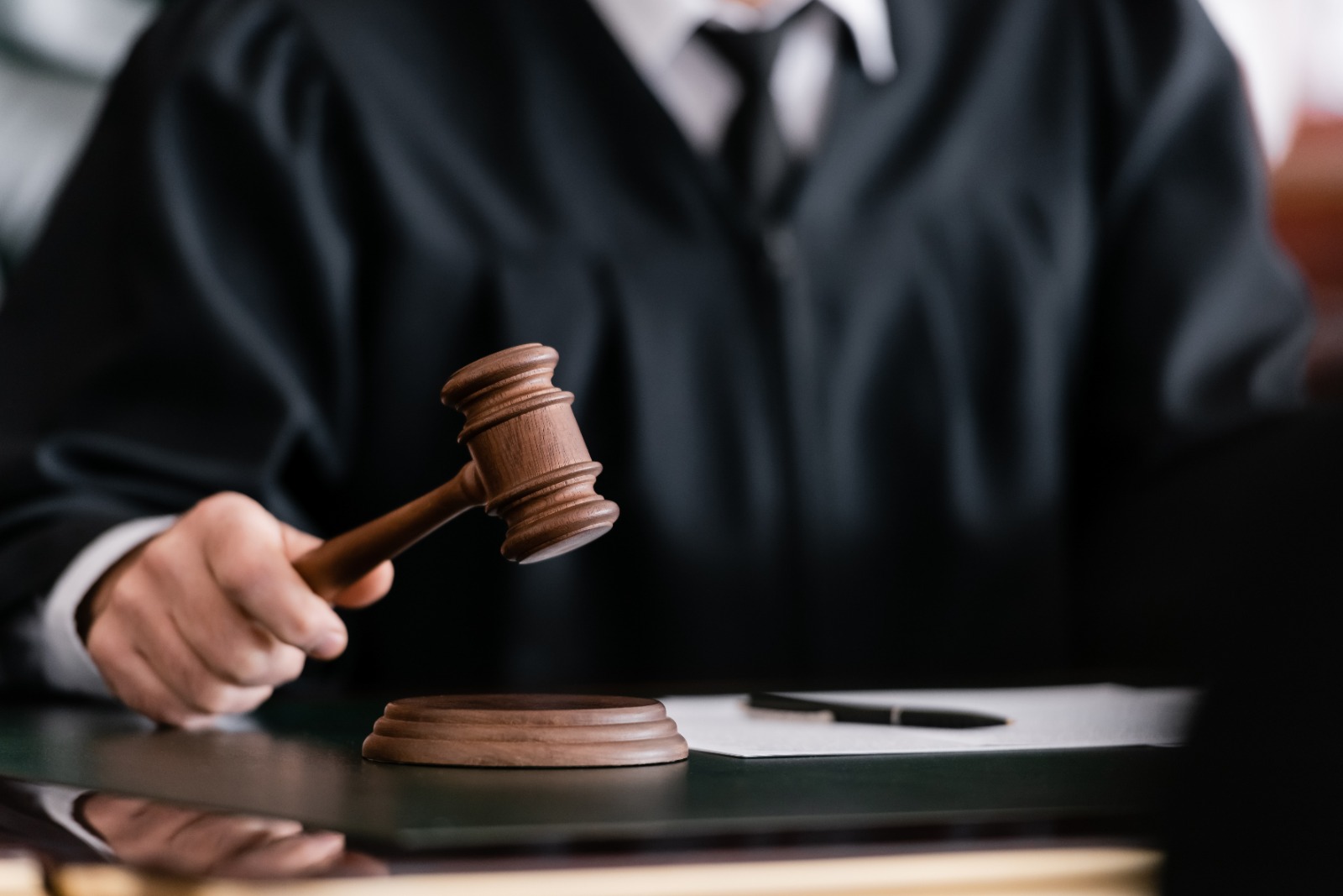 Imagem mostra um homem vestido com capa preta, camisa social branca por dentro e gravata preta, segurando um martelo de Justiça