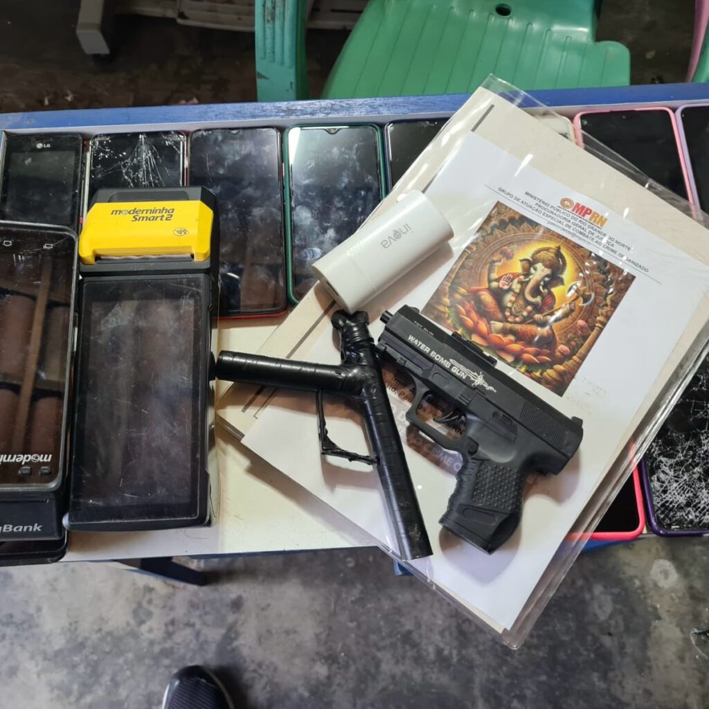 Imagem de vários celulares, armas e maquinetas de cartão arrumados em cima de uma mesa.