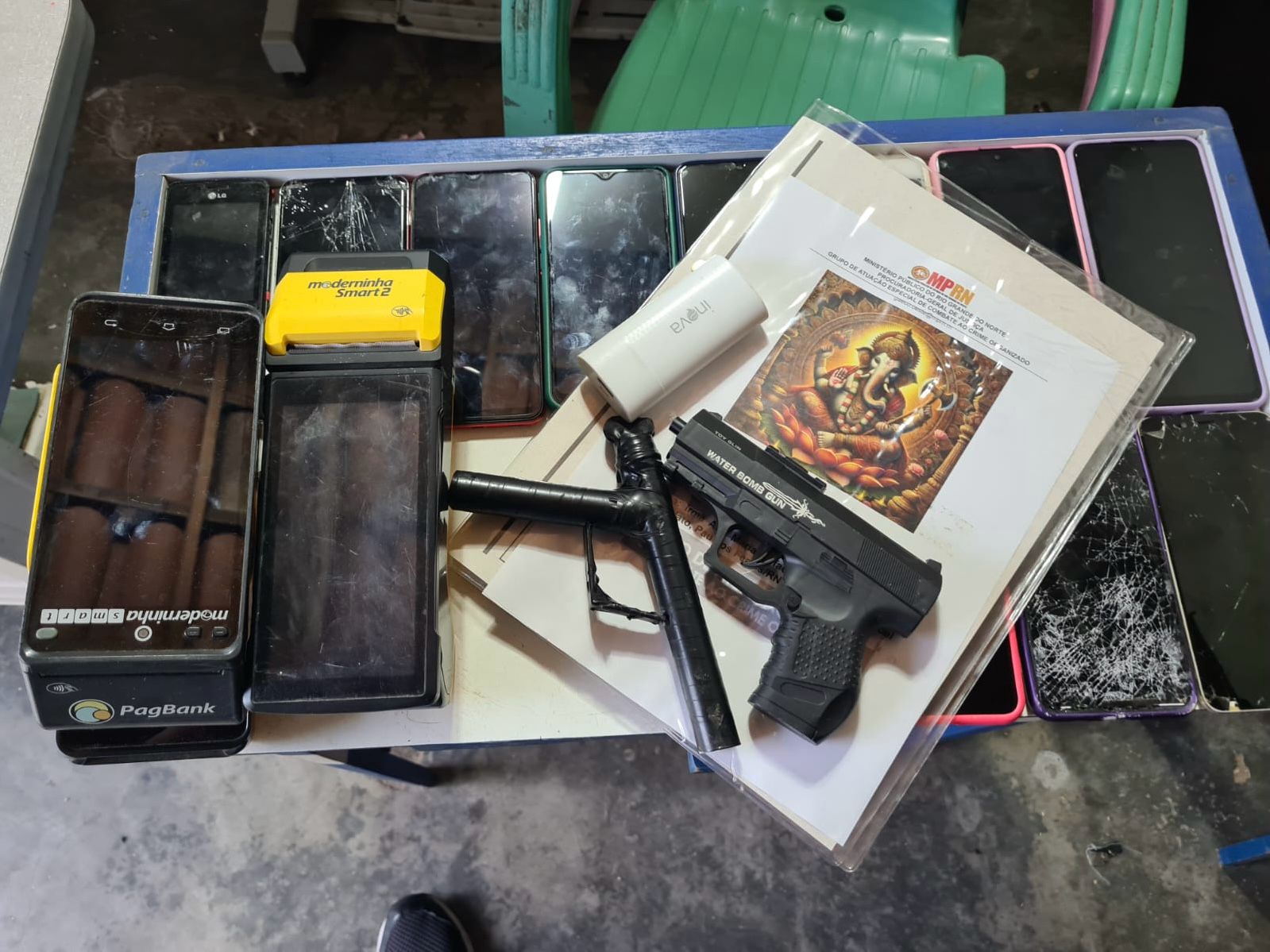 Imagem de vários celulares, armas e maquinetas de cartão arrumados em cima de uma mesa.