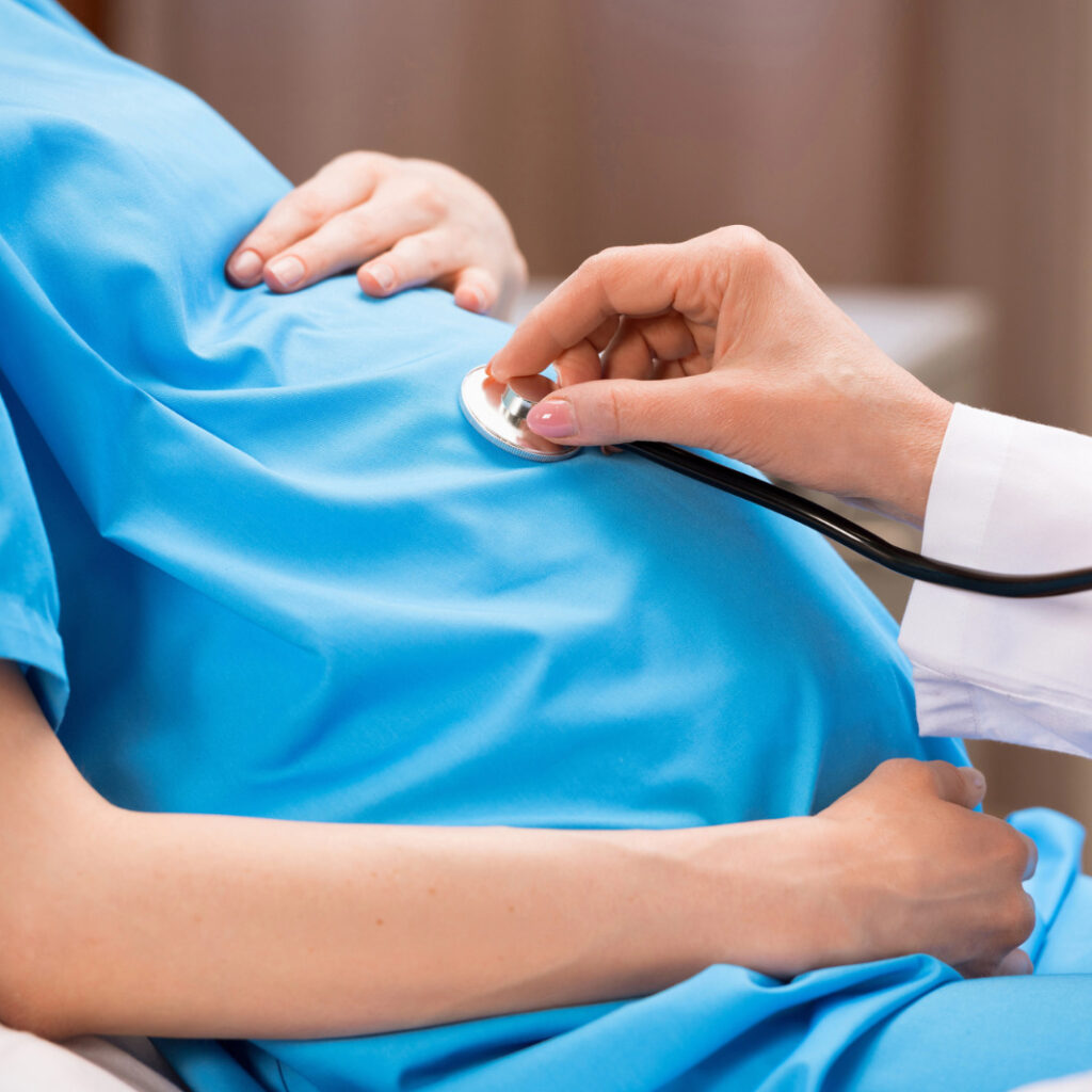 imagem de uma mulher gravida vestindo uma bata azul e uma medico com estetoscopio na barriga dela