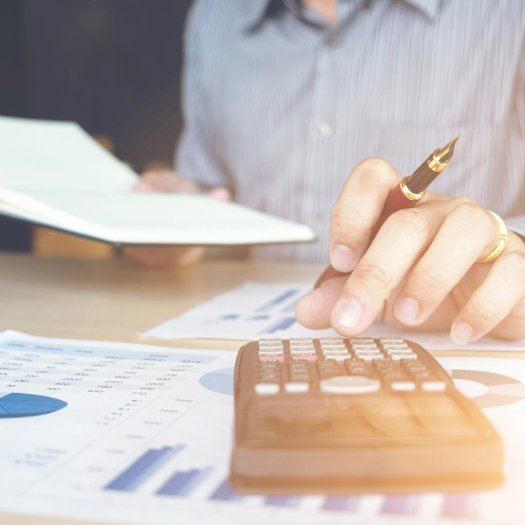 Homem usando uma calculadora e segurando uma caneta sobre uma mesa com relatórios financeiros e um caderno aberto.