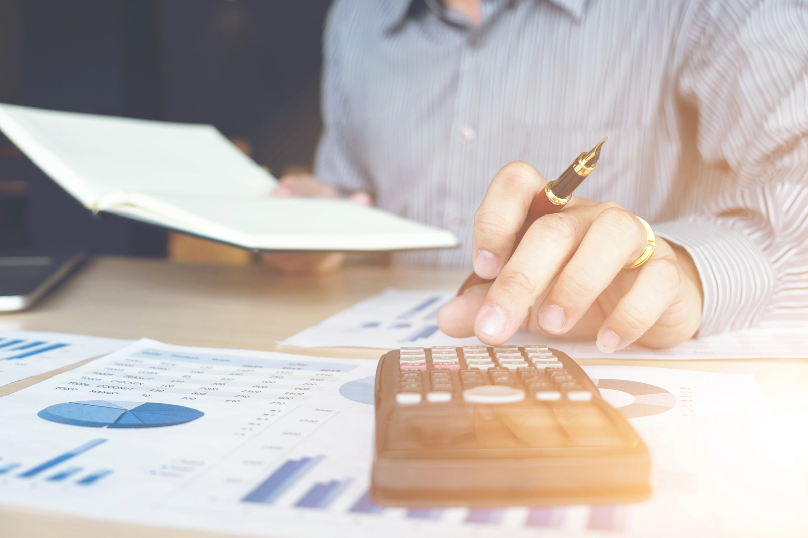 Homem usando uma calculadora e segurando uma caneta sobre uma mesa com relatórios financeiros e um caderno aberto.