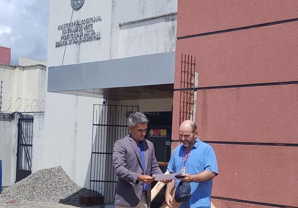 Dois homens seguram e analisam um documento em frente ao prédio do Ministério Público do Rio Grande do Norte.