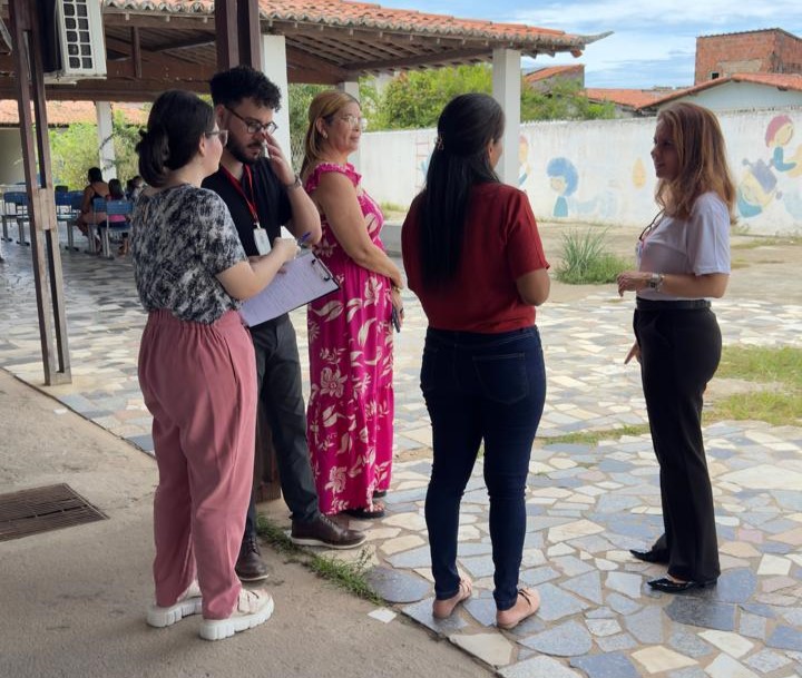 imagem mostra pessoas conversando em um pátio aberto, com uma mulher à direita de frente para o grupo enquanto um homem à esquerda faz anotações em uma prancheta.