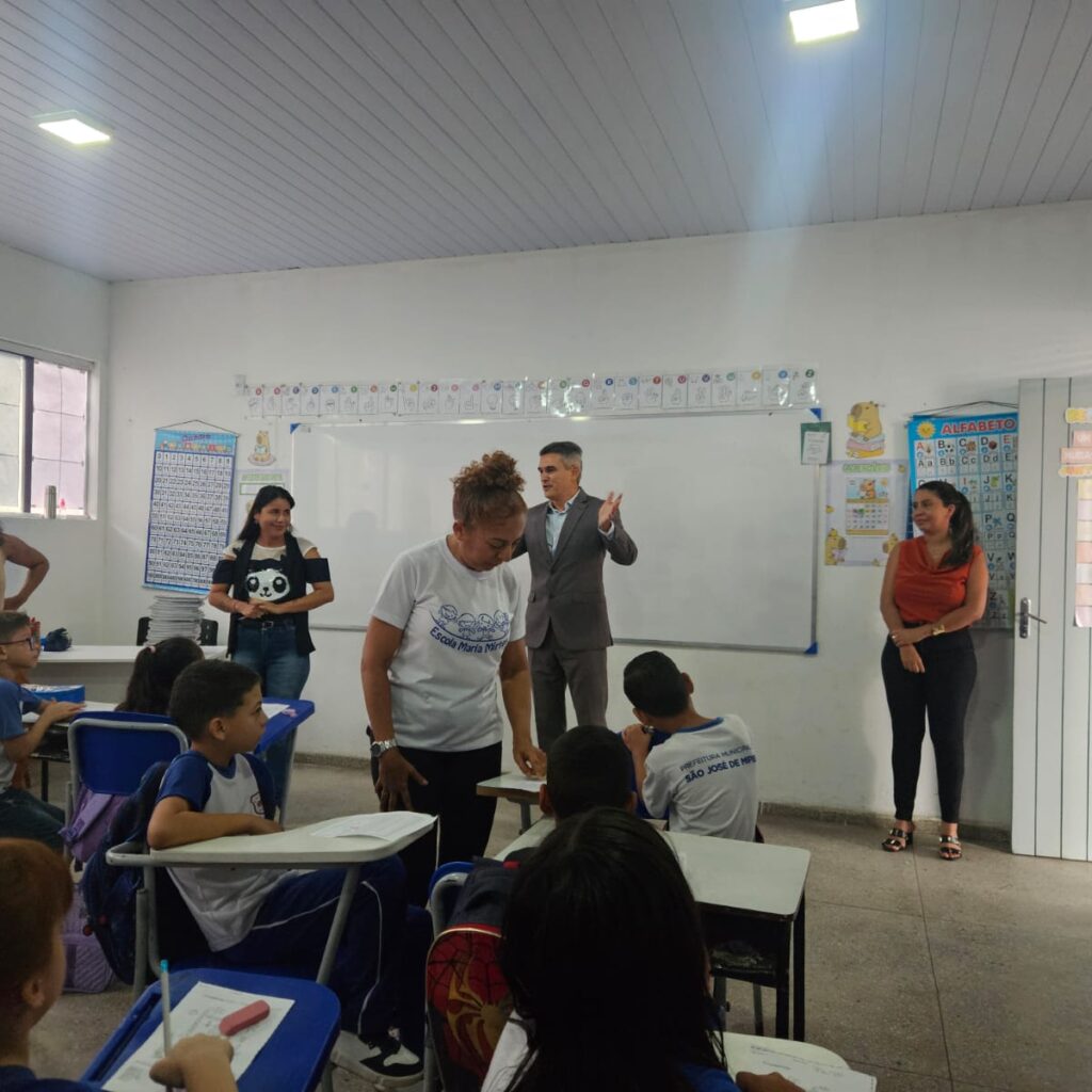 Interior de uma sala de aula com alunos sentados em suas bancas enquanto adultos, incluindo um homem de terno gesticulando à frente do quadro branco, interagem durante uma atividade escolar.
