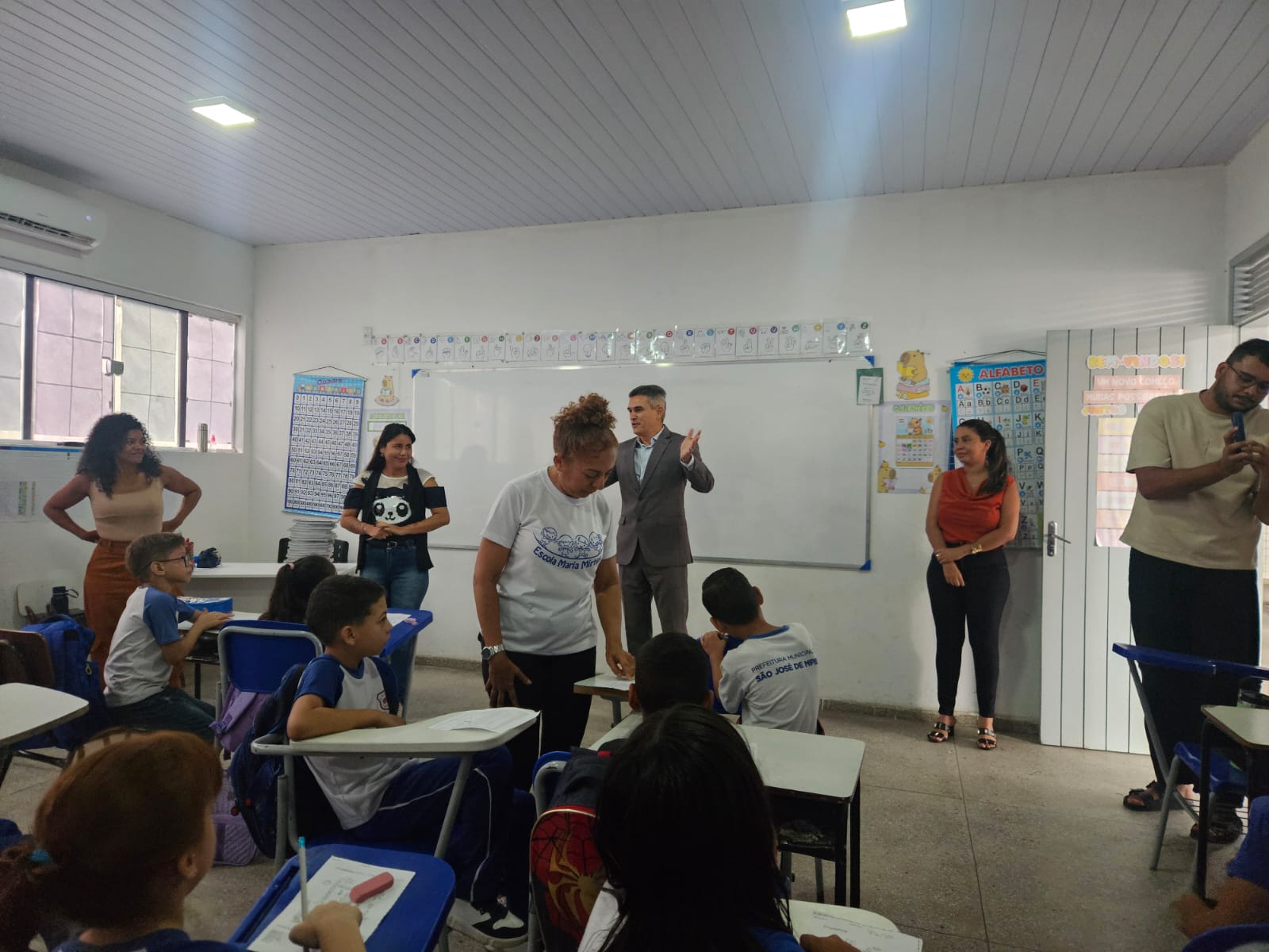 Interior de uma sala de aula com alunos sentados em suas bancas enquanto adultos, incluindo um homem de terno gesticulando à frente do quadro branco, interagem durante uma atividade escolar.