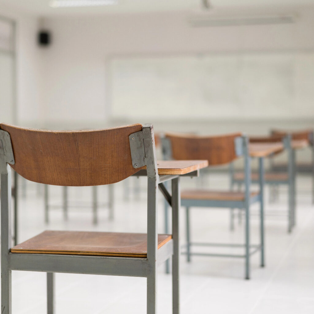 Uma sala de aula vazia com foco em uma cadeira escolar de madeira e metal em primeiro plano, com outras fileiras de carteiras e um quadro branco ao fundo de forma desfocada.
