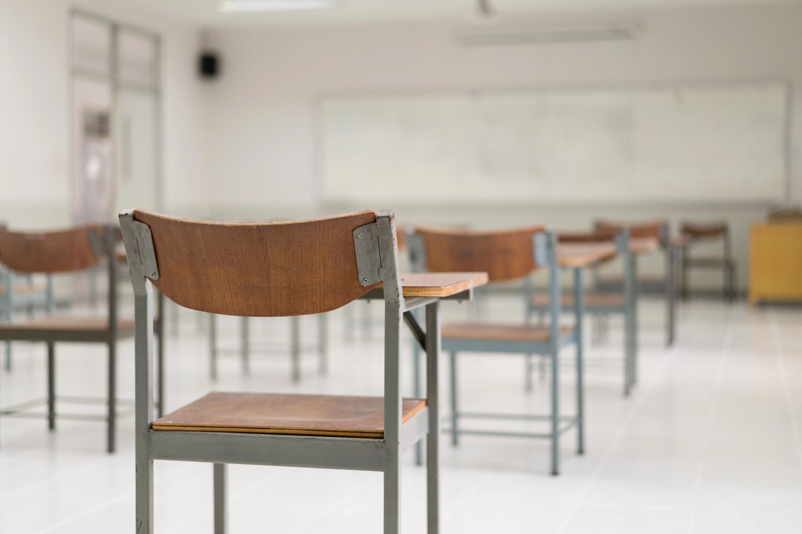Uma sala de aula vazia com foco em uma cadeira escolar de madeira e metal em primeiro plano, com outras fileiras de carteiras e um quadro branco ao fundo de forma desfocada.