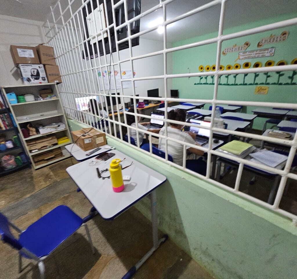A imagem mostra o interior de uma sala de aula ou de estudos, separada por uma grade branca em primeiro plano. Fora da grade, há uma mesa com uma cadeira azul e uma garrafa de água amarela e rosa. No interior da sala, alunos estão sentados em mesas, mexendo em computadores. Ao fundo, uma estante com livros e decorações de girassol na parede.