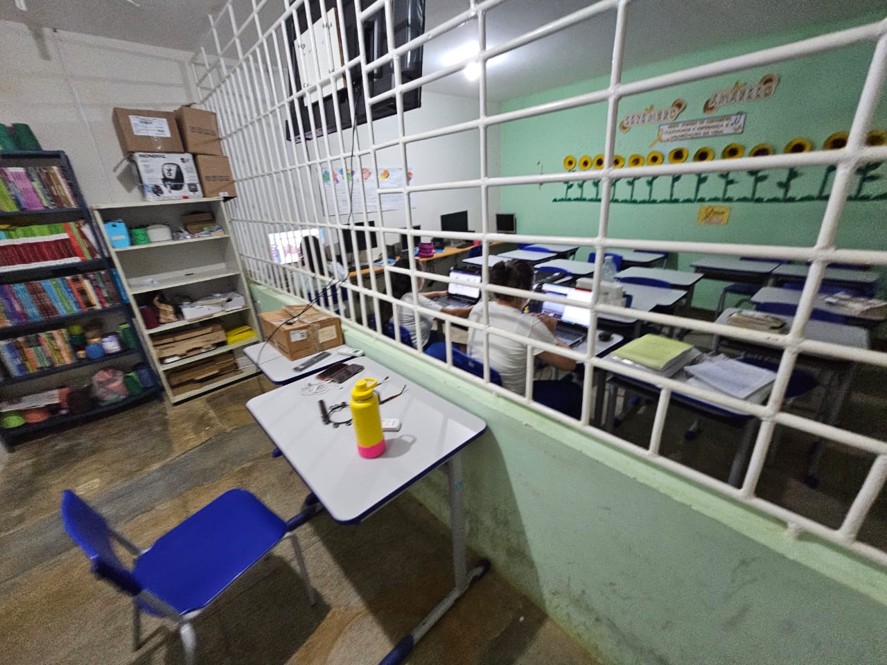 A imagem mostra o interior de uma sala de aula ou de estudos, separada por uma grade branca em primeiro plano. Fora da grade, há uma mesa com uma cadeira azul e uma garrafa de água amarela e rosa. No interior da sala, alunos estão sentados em mesas, mexendo em computadores. Ao fundo, uma estante com livros e decorações de girassol na parede.