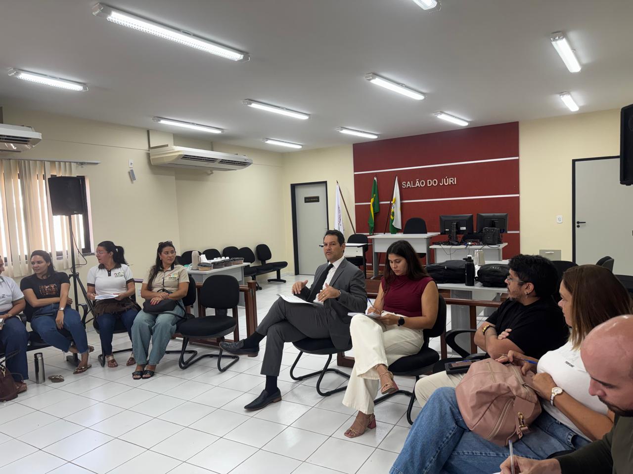 Pessoas sentadas em círculo participando de uma reunião em uma sala com a inscrição "Salão do Júri" ao fundo e o promoto de justica ao centro gesticulando enquanto fala