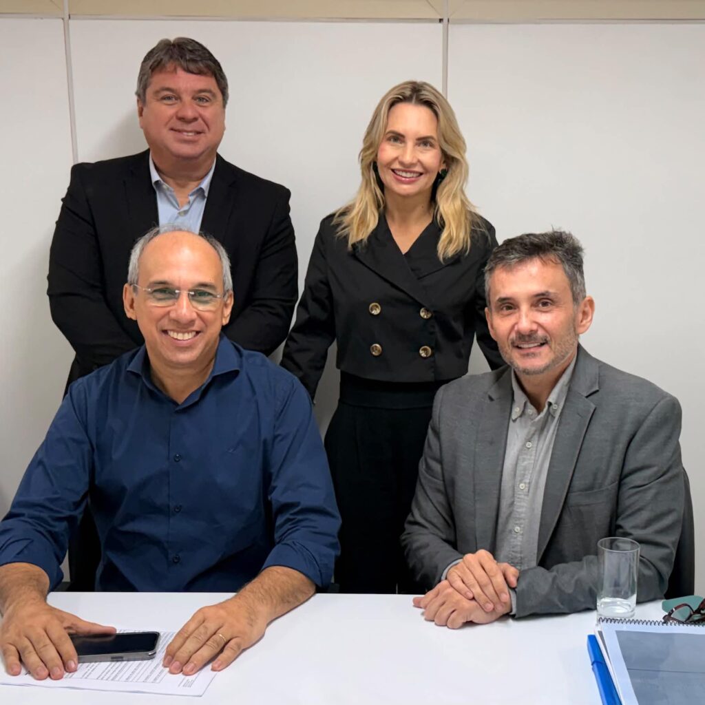 Quatro pessoas, sendo três homens e uma mulher, sorriem para a foto em um ambiente de escritório, com dois homens sentados à mesa e o outro casal em pé logo atrás.