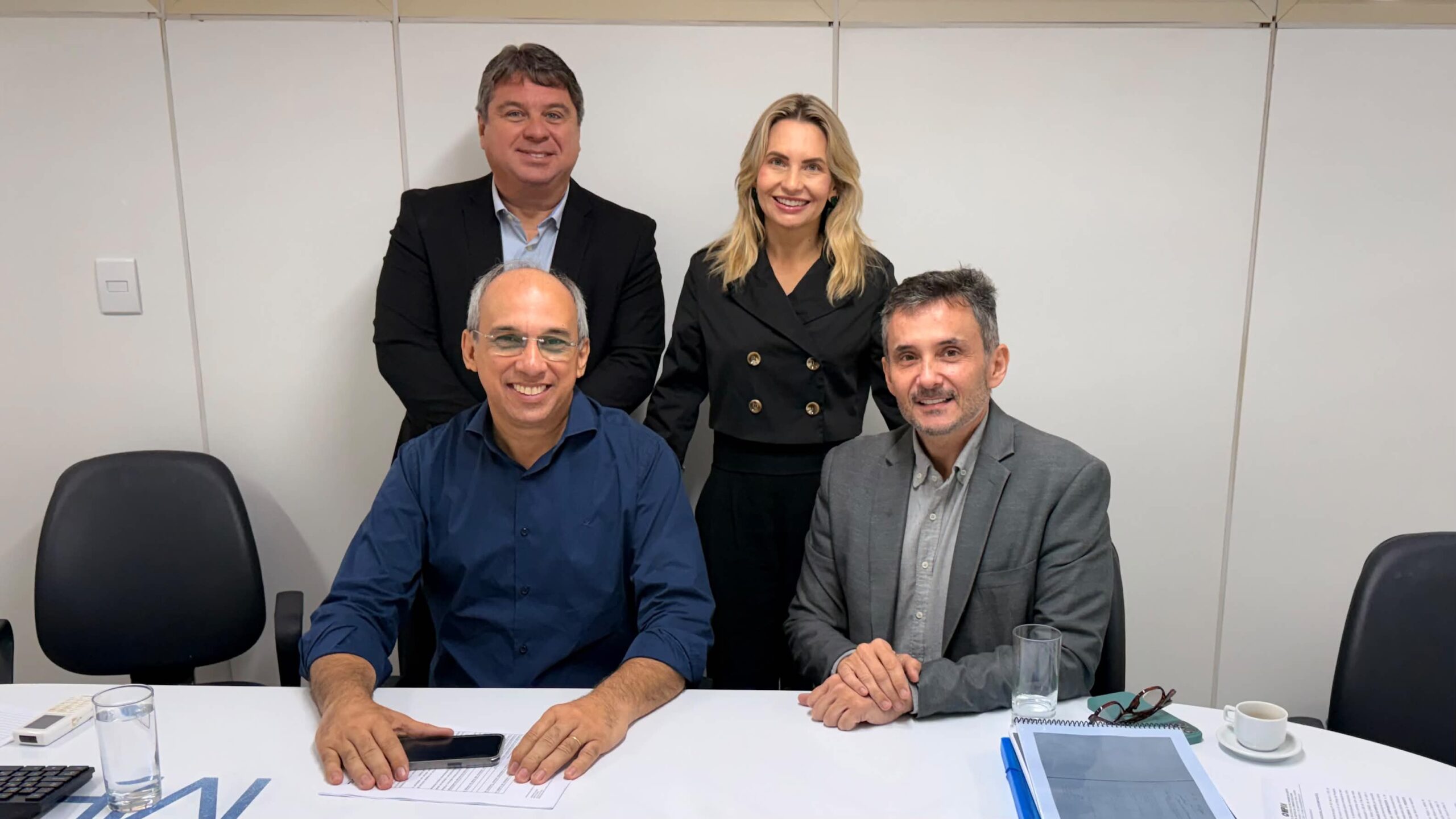 Quatro pessoas, sendo três homens e uma mulher, sorriem para a foto em um ambiente de escritório, com dois homens sentados à mesa e o outro casal em pé logo atrás.