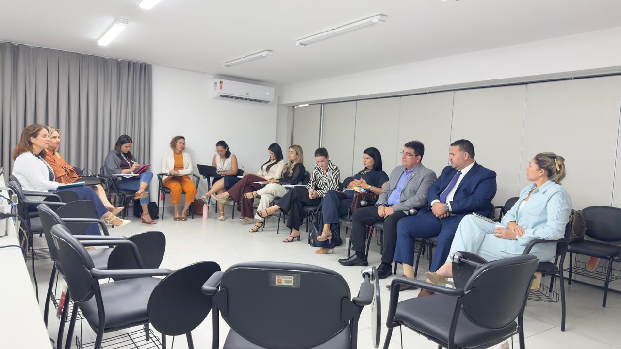 Uma reunião com cerca de doze pessoas sentadas em círculo em uma sala de paredes brancas, vestindo trajes formais e casuais enquanto conversam e fazem anotações