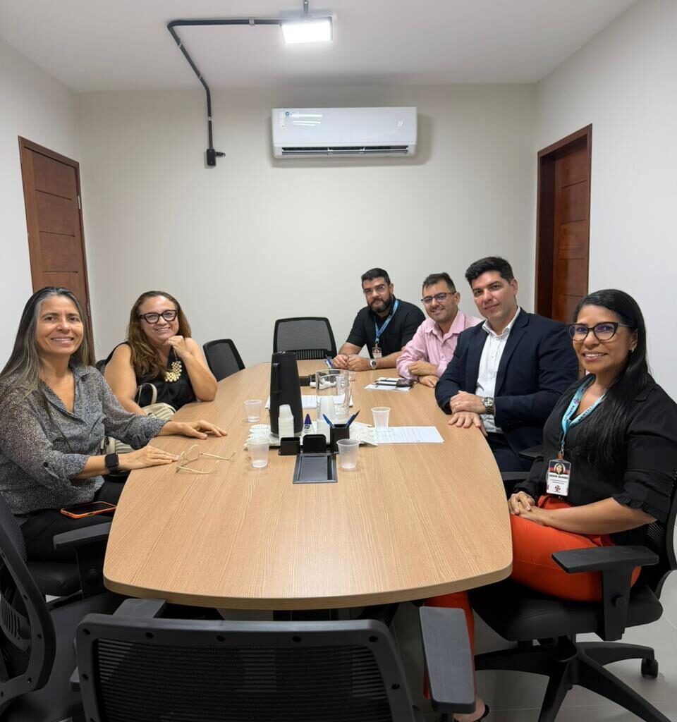 Seis pessoas, sendo três mulheres e três homens, estão sentadas sorrindo ao redor de uma grande mesa de reunião de madeira em um escritório com paredes brancas.